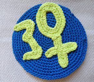 Gehäkeltes BF-Logo: Eine gelbe 30, statt 0 ein Venussymbol auf blauem gehäkeltem Grund
